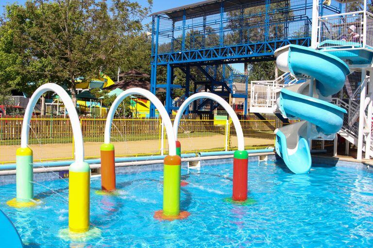 Arcos de colores que lanzan agua en la piscina Acuaspray, con los toboganes al fondo